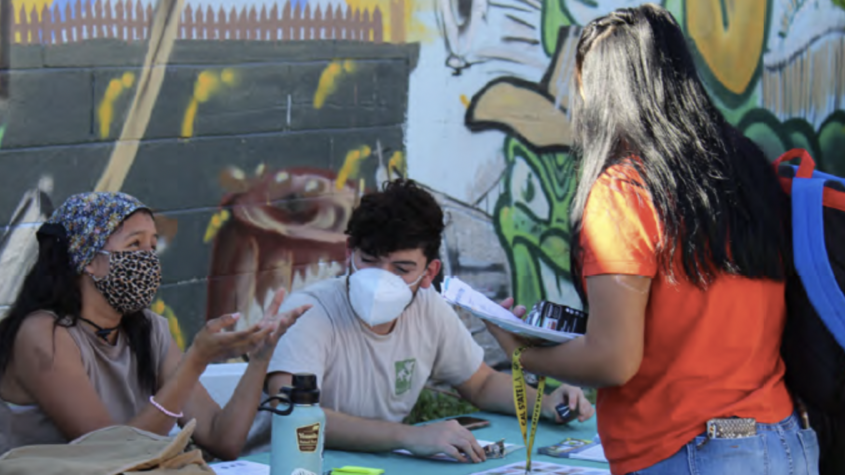 A woman in an orange shirt talks to a man and a woman wearing masks and sitting at a booth in front of a mural at the Healthy Watts Harvest community engagement event in November 2021