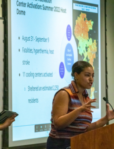 woman in striped top speaks at podium while map is projected on a screen behind her